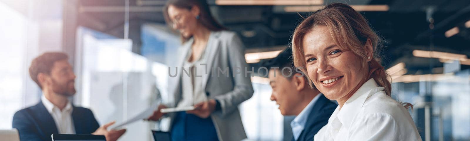 confident smiling businesswoman sitting at the office with group of colleagues, on background by Yaroslav_astakhov