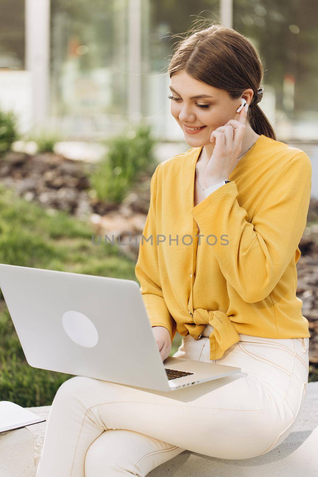 Portrait of smiling business woman looking at laptop screen. Focused businesswoman using wireless earphones working on laptop computer near modern office. Female manager typing on laptop keyboard.