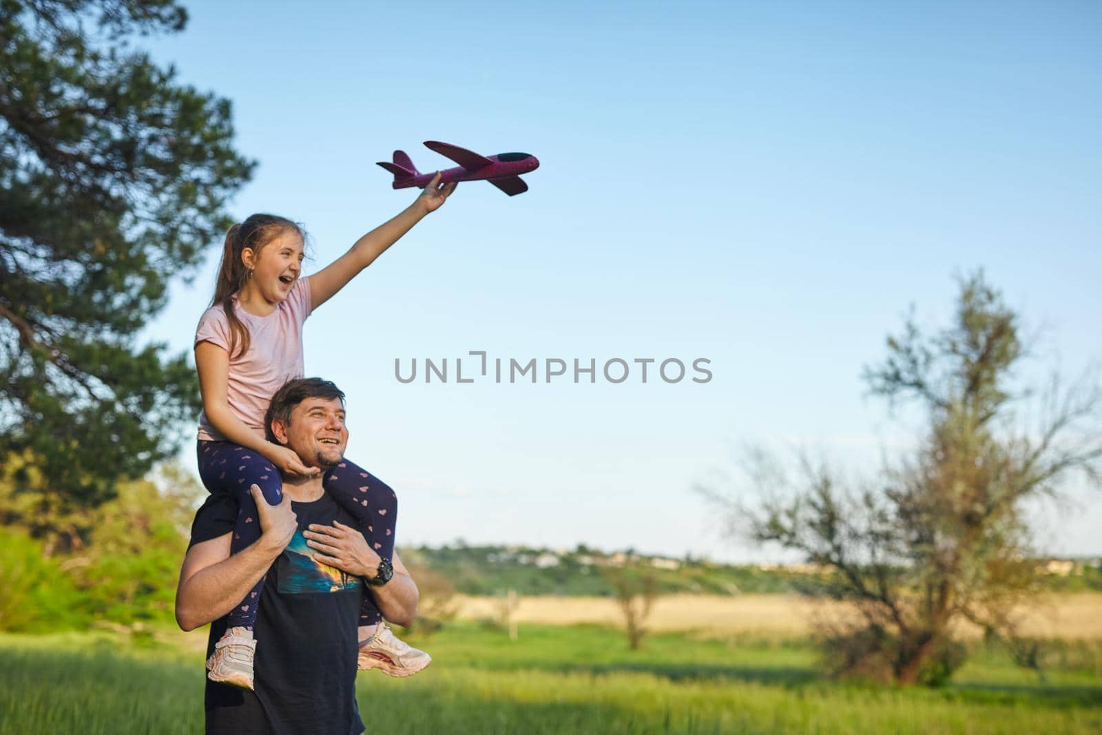 Cute girl riding on father's shoulder and playing with toy airplane against sky in summer forest