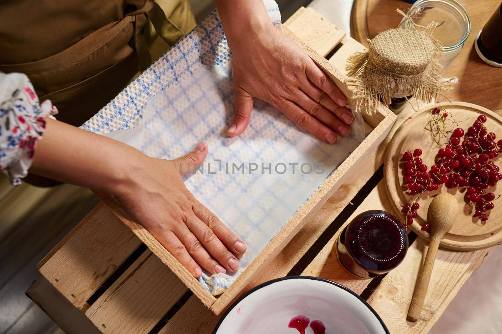 Details: Hands of a woman housewife arranging jars with handmade jam upside down in a wooden crate and covering them with a waffle towel. Red currant berries lying down on wooden board