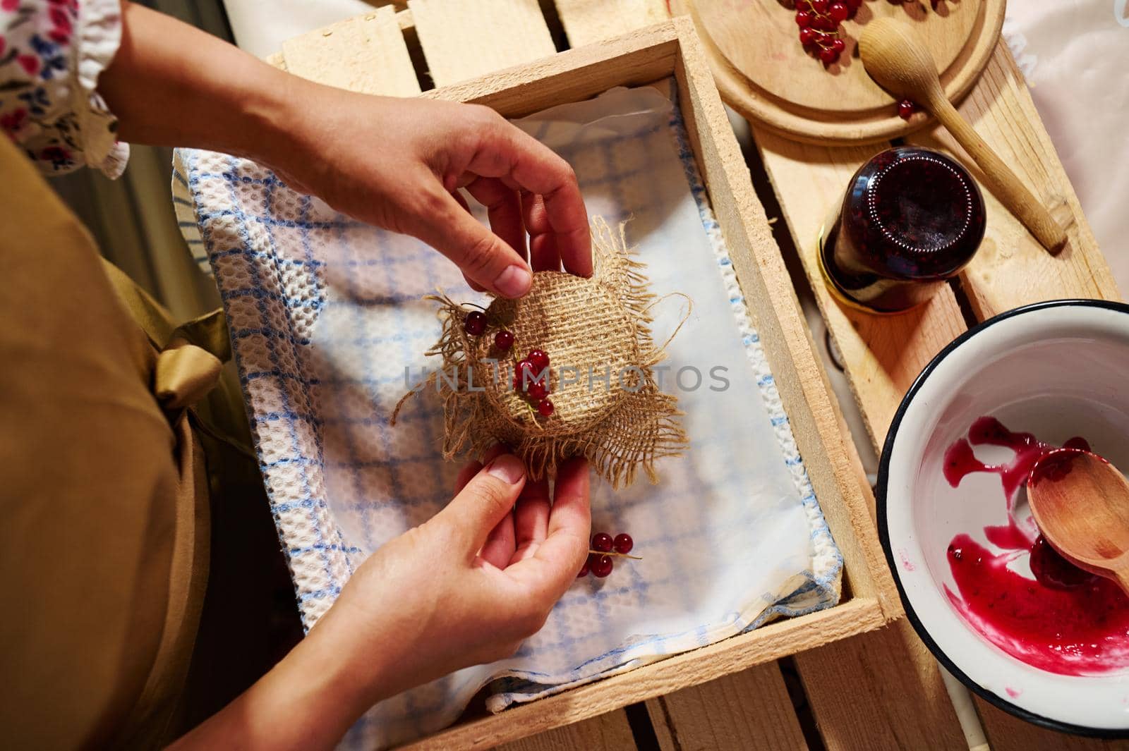 Preparing preserves, marmalades, confitures and jelly at home kitchen. Close-up hands of a housewife tying bow on the burlap on a cover, decorating a jar of homemade red currant berry jam.