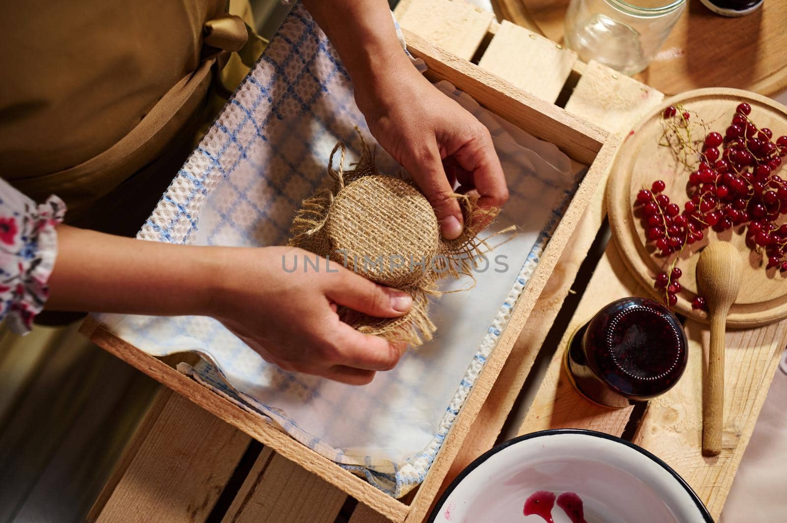 Woman hands putting burlap on the lid of a jar with homemade red currant jelly, tying a bow of rope. Berries lying on a wooden board next to kitchen spoon, bowl and jar of jam upside down on a crate