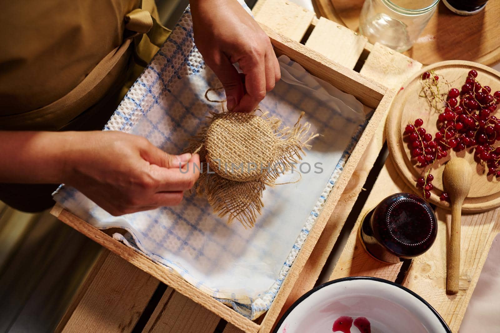 Top view. Housewife ties bow on the burlap of a lid, ornates a jar of homemade delicious jelly from red currant berries by artgf