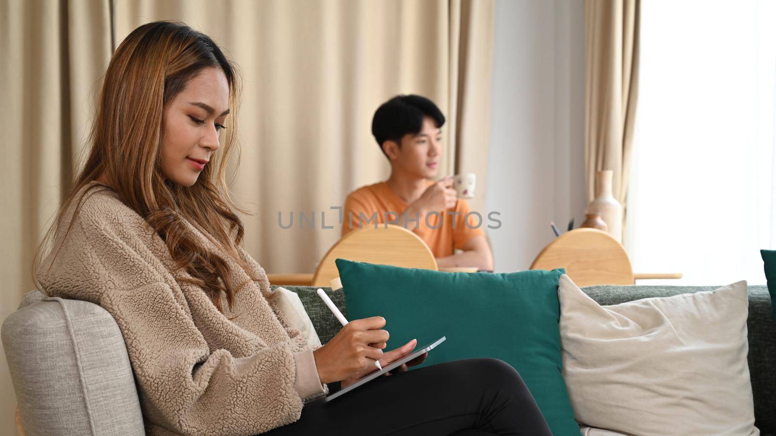 Young asian woman using digital tablet on sofa and her husband sitting in background. by prathanchorruangsak