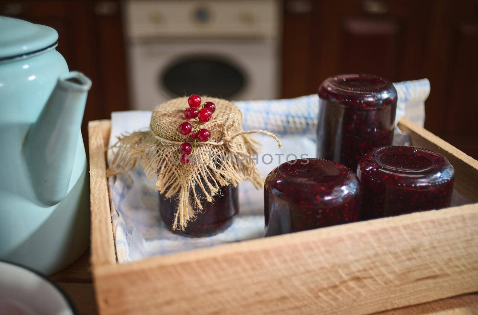 Still life. Jars with homemade red currant jam on wooden crate and vintage blue enamel teapot on kitchen table by artgf