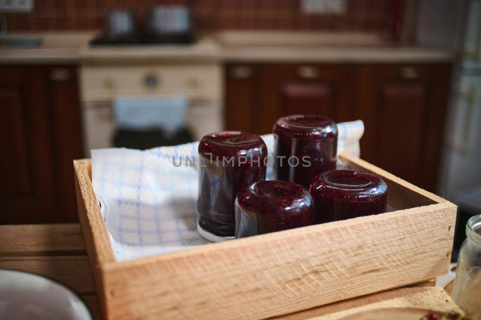 Focus on jars with berry jam stacking upside down on a wooden crate, standing on kitchen island in the home rustic cozy kitchen interior. Canned and preserved food for winter season