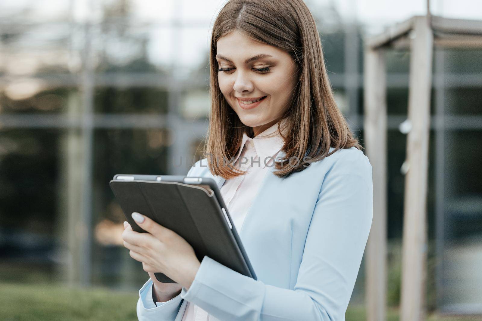 Businesswoman holding tablet in hands using business apps on tablet computer near office building. Happy face looking tablet screen outdoors. Elegant female chatting online with digital device outside