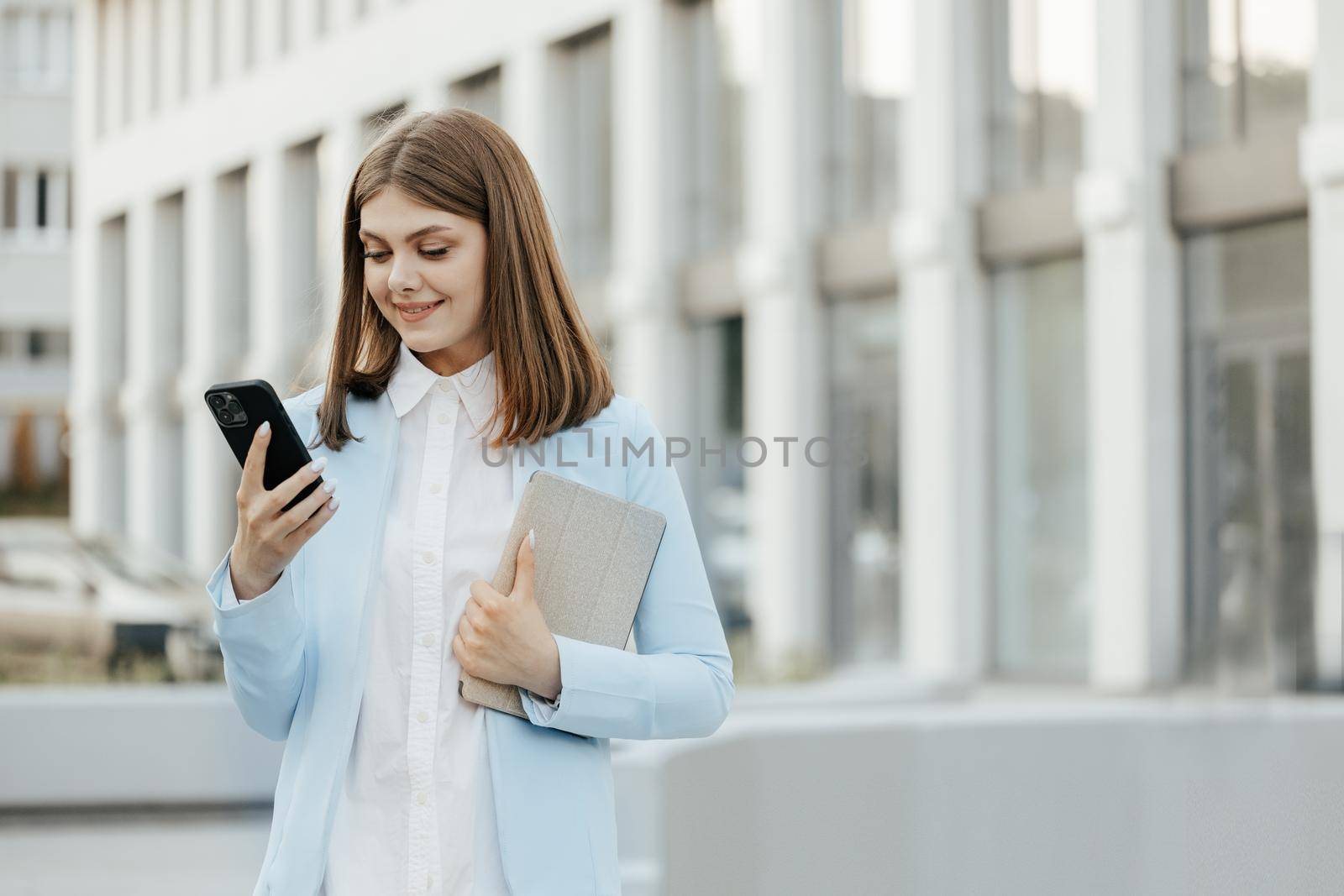 Smiling caucasian woman wearing formal suit using mobile phone typing text messages walking holding tablet computer outside business cellphone city smartphone. by uflypro