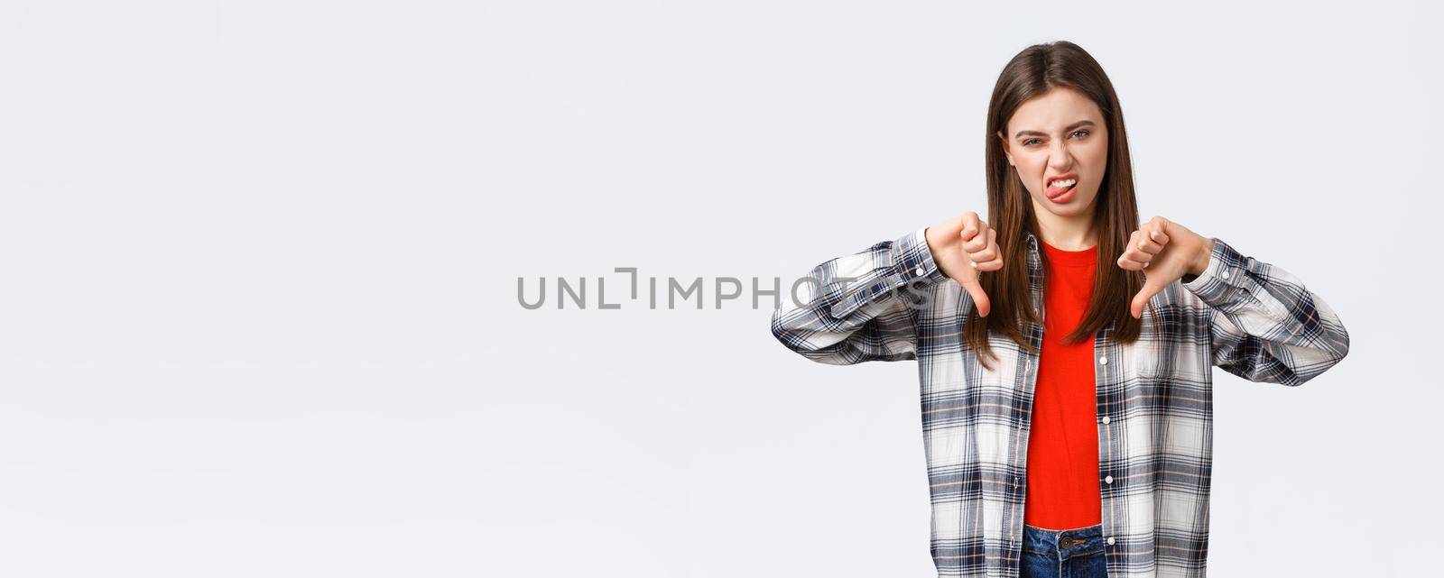 Lifestyle, different emotions, leisure activities concept. Displeased and unimpressed young picky girl in checked shirt, thumb-down and show tongue to express dislike, rate awful movie by Benzoix