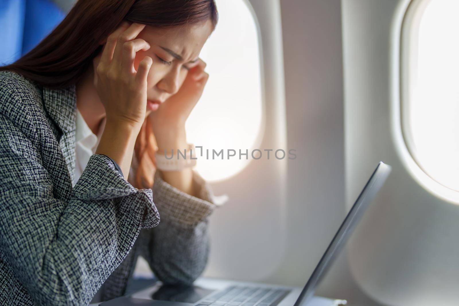 portrait of a successful Asian businesswoman or entrepreneur in a formal suit on an airplane seated in Business Class shows a thoughtful and stressed face with using laptop during the flight.