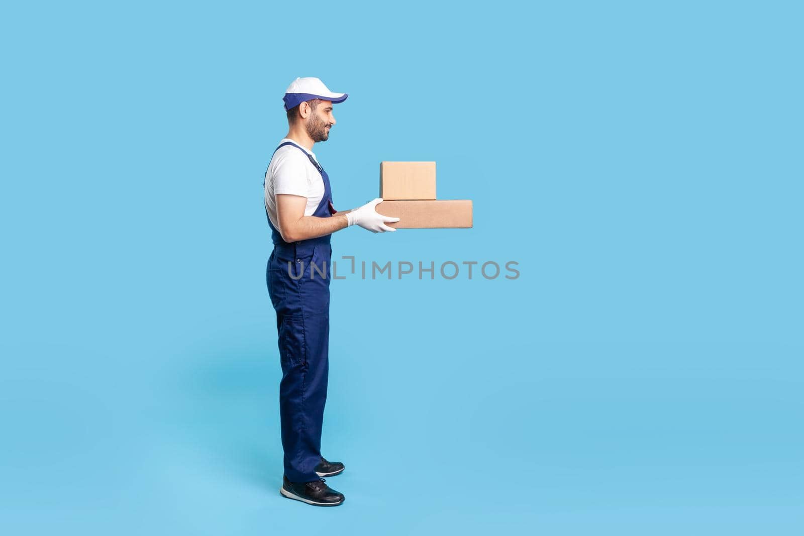 Side view expert delivery man in uniform with gloves giving cardboard boxes to client and smiling, carrying ordered purchase. Profession of courier, shipment and cargo transportation service. isolated