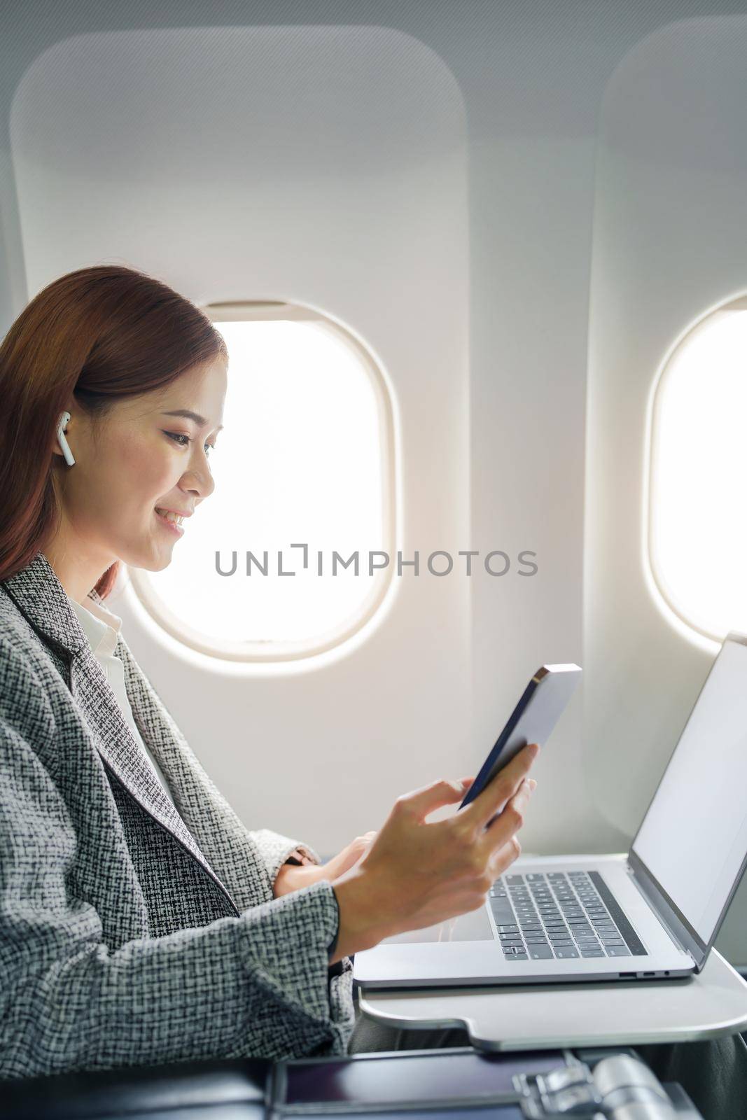 A successful asian businesswoman or female entrepreneur in formal suit in a plane sits in a business class's seat and uses a smartphone during flight by Manastrong