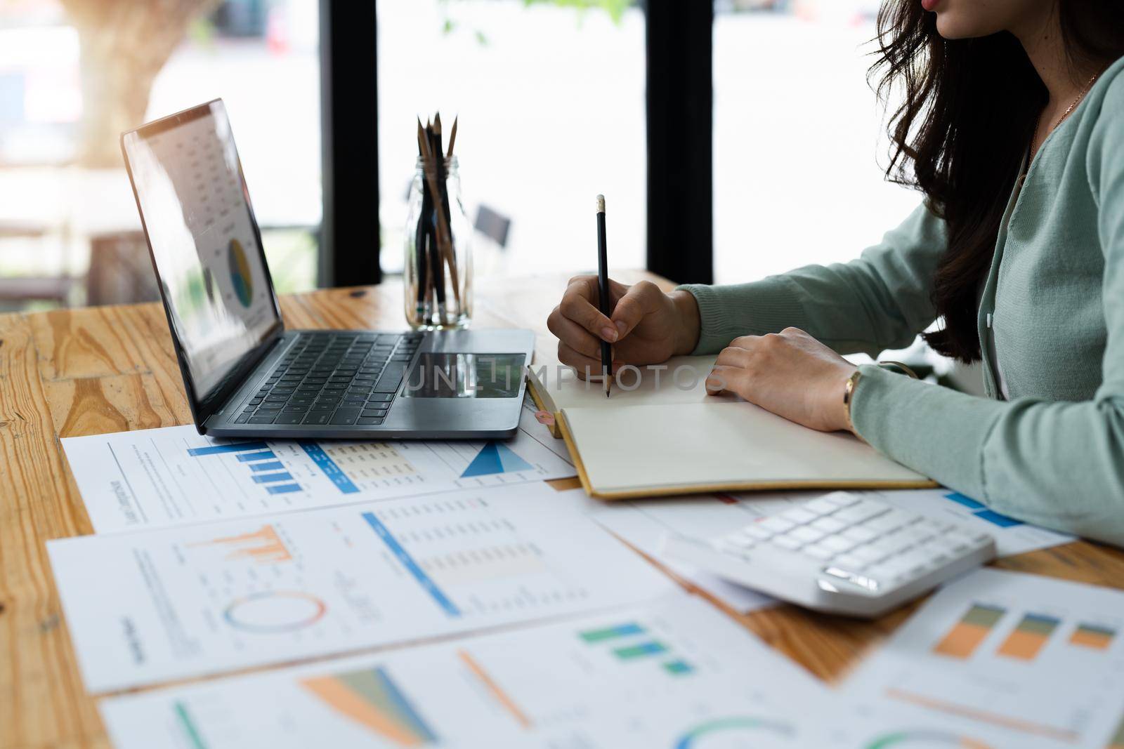 businesswoman or accountant taking note and working on calculator and laptop computer to calculate business data during make note at notepad, accountancy document at office. by nateemee