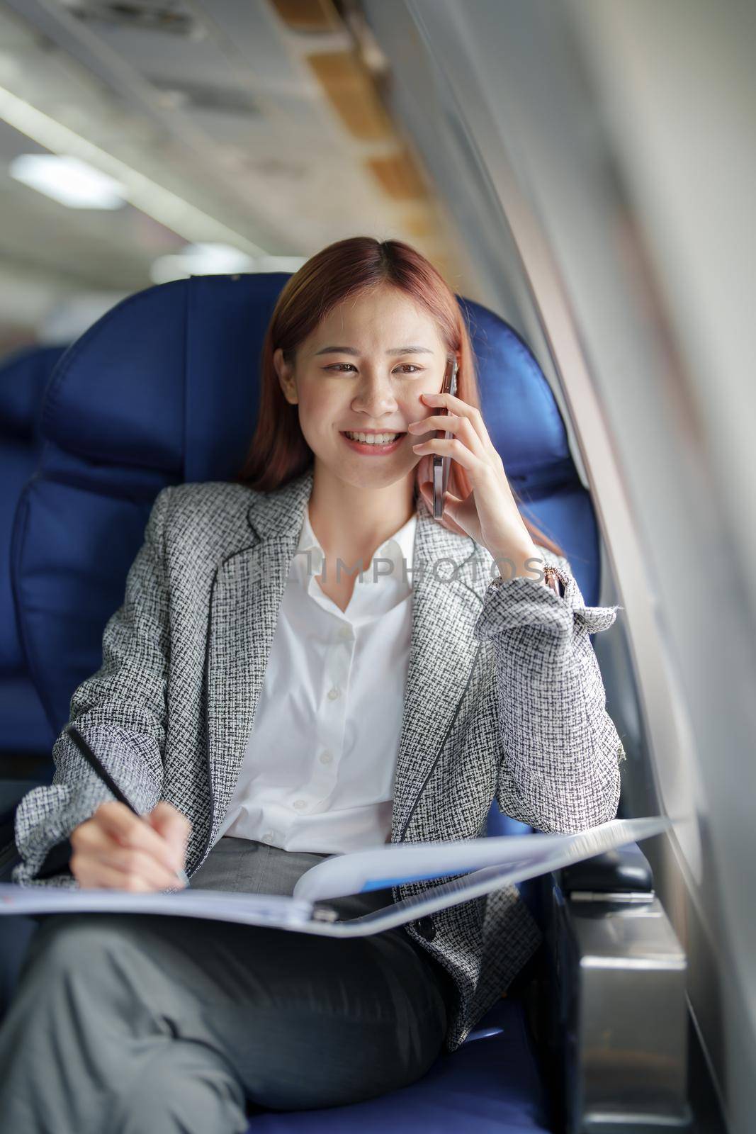 portrait of A successful asian business woman or female entrepreneur in formal suit in a plane sits in a business class seat and uses smartphone with documents for work during flight.