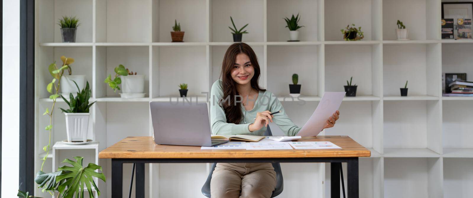 Portrait of happy Asian businesswoman using laptop computer while working with using a calculator to calculate the numbers, finance accounting concept - banner shot.