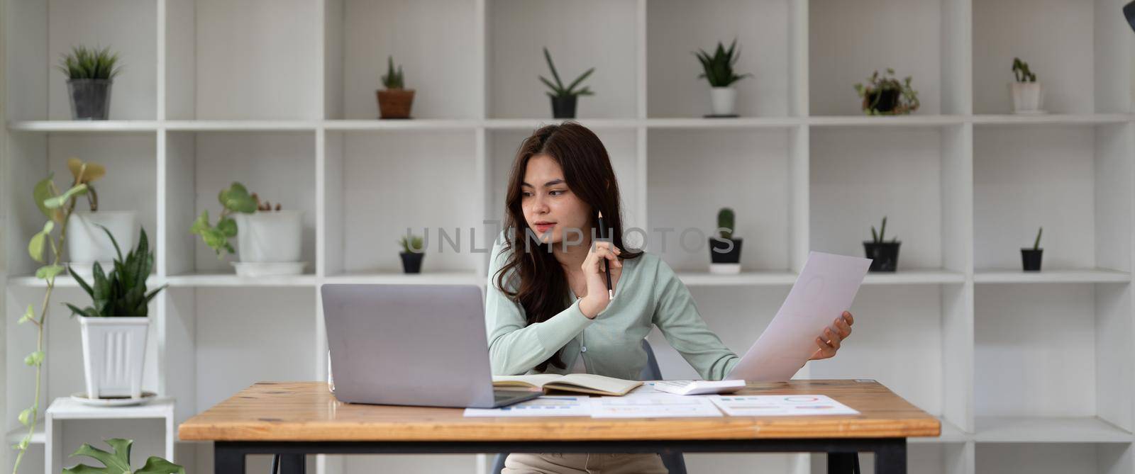 Portrait of happy Asian businesswoman using laptop computer while working with using a calculator to calculate the numbers, finance accounting concept - banner shot.