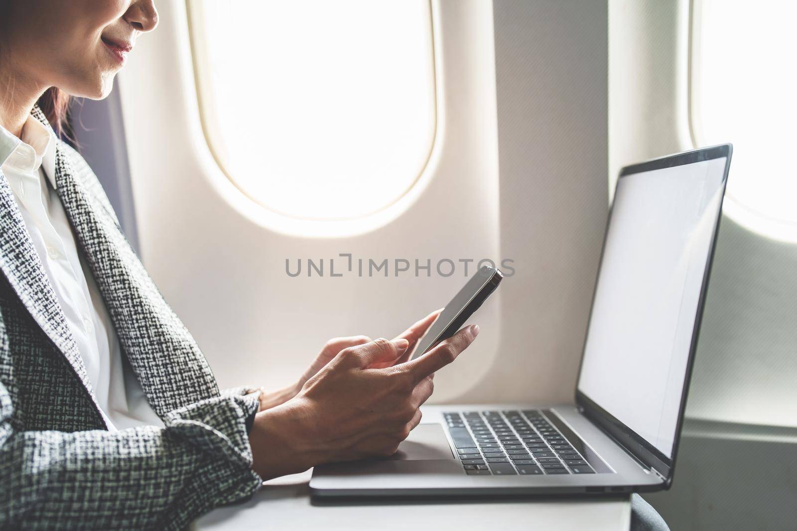 A successful asian businesswoman or female entrepreneur in formal suit in a plane sits in a business class's seat using a smartphone and computer laptop during flight.