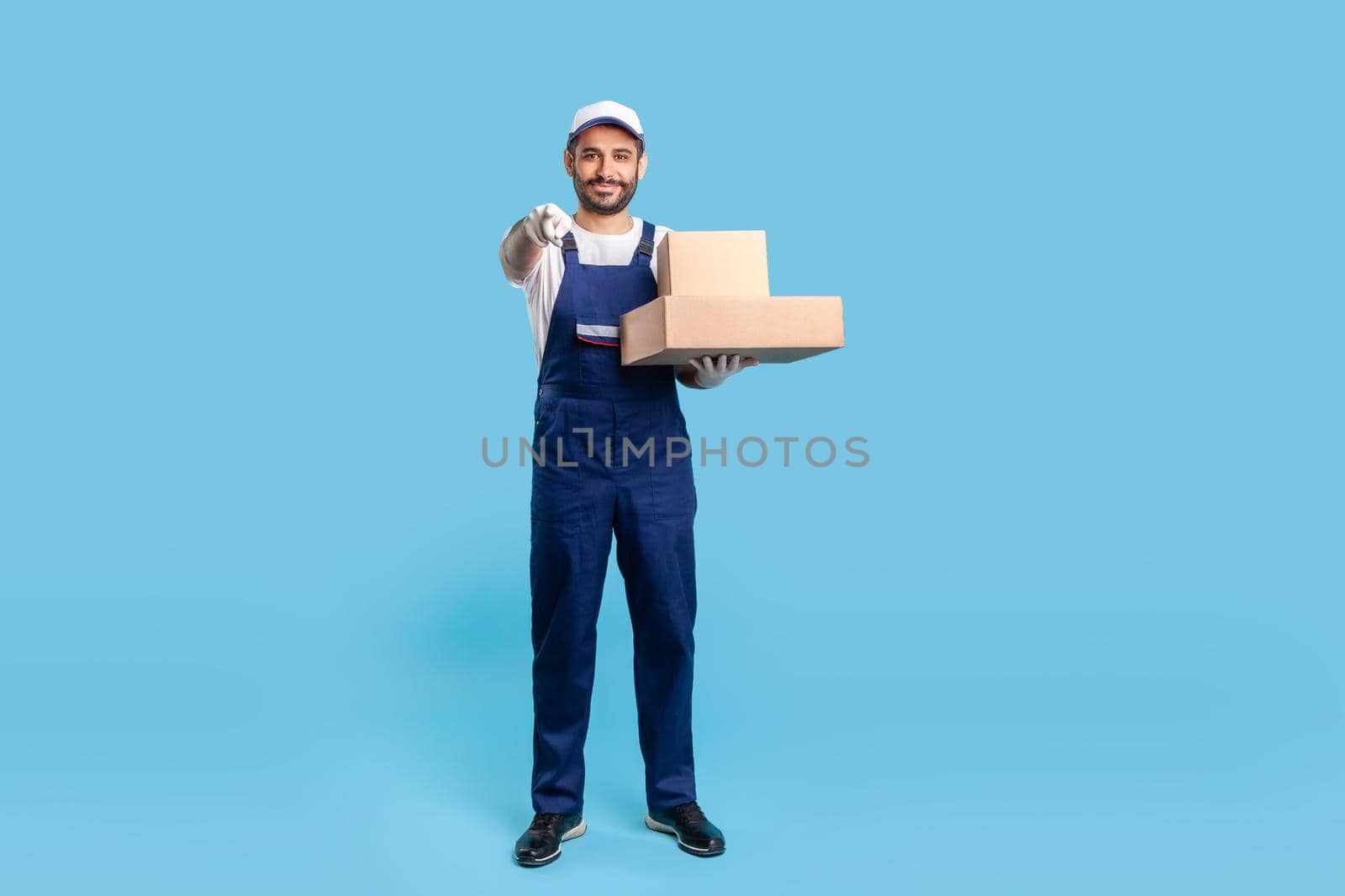 Full length professional delivery man in uniform with gloves holding cardboard boxes and pointing to camera, choosing you. Profession of courier, shipment and cargo transportation service. isolated