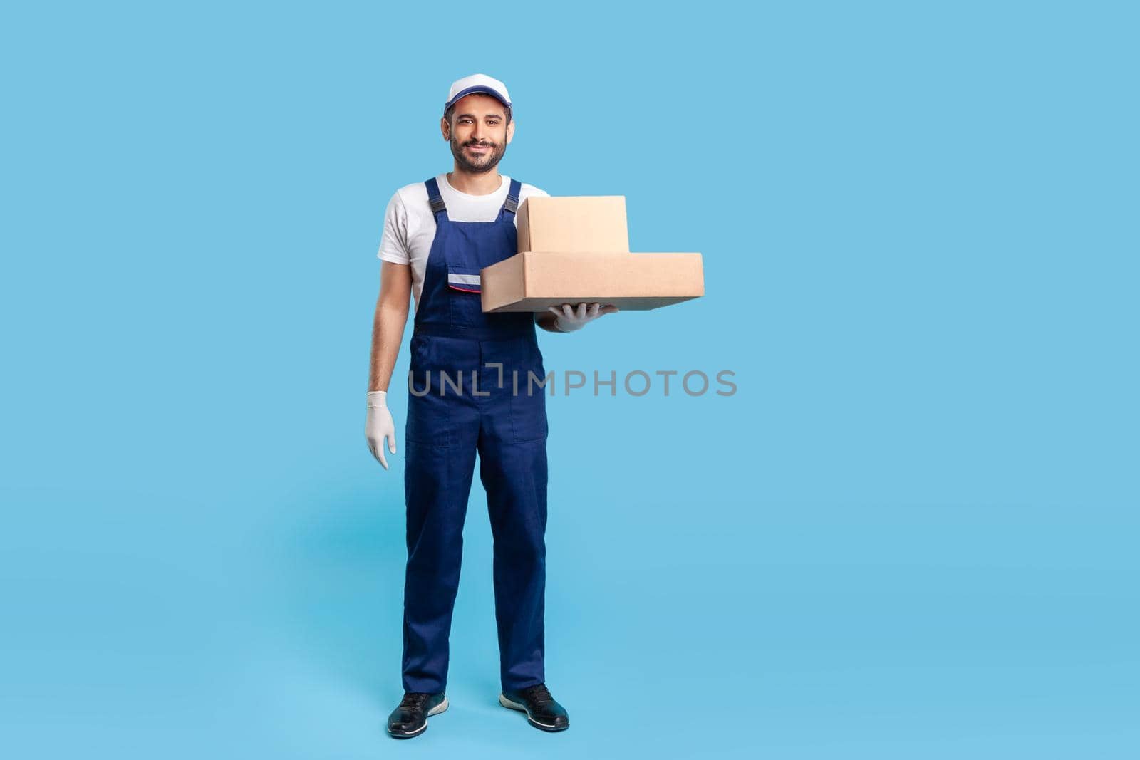 Full length delivery man in blue uniform with gloves standing, holding cardboard boxes and smiling to camera. Profession of courier, shipment and cargo transportation service. studio shot isolated