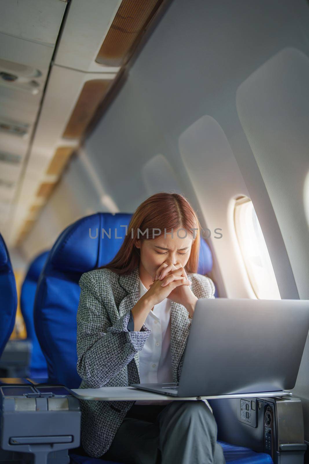 portrait of a successful Asian businesswoman or entrepreneur in a formal suit on an airplane seated in Business Class shows a thoughtful and stressed face with using laptop during the flight by Manastrong