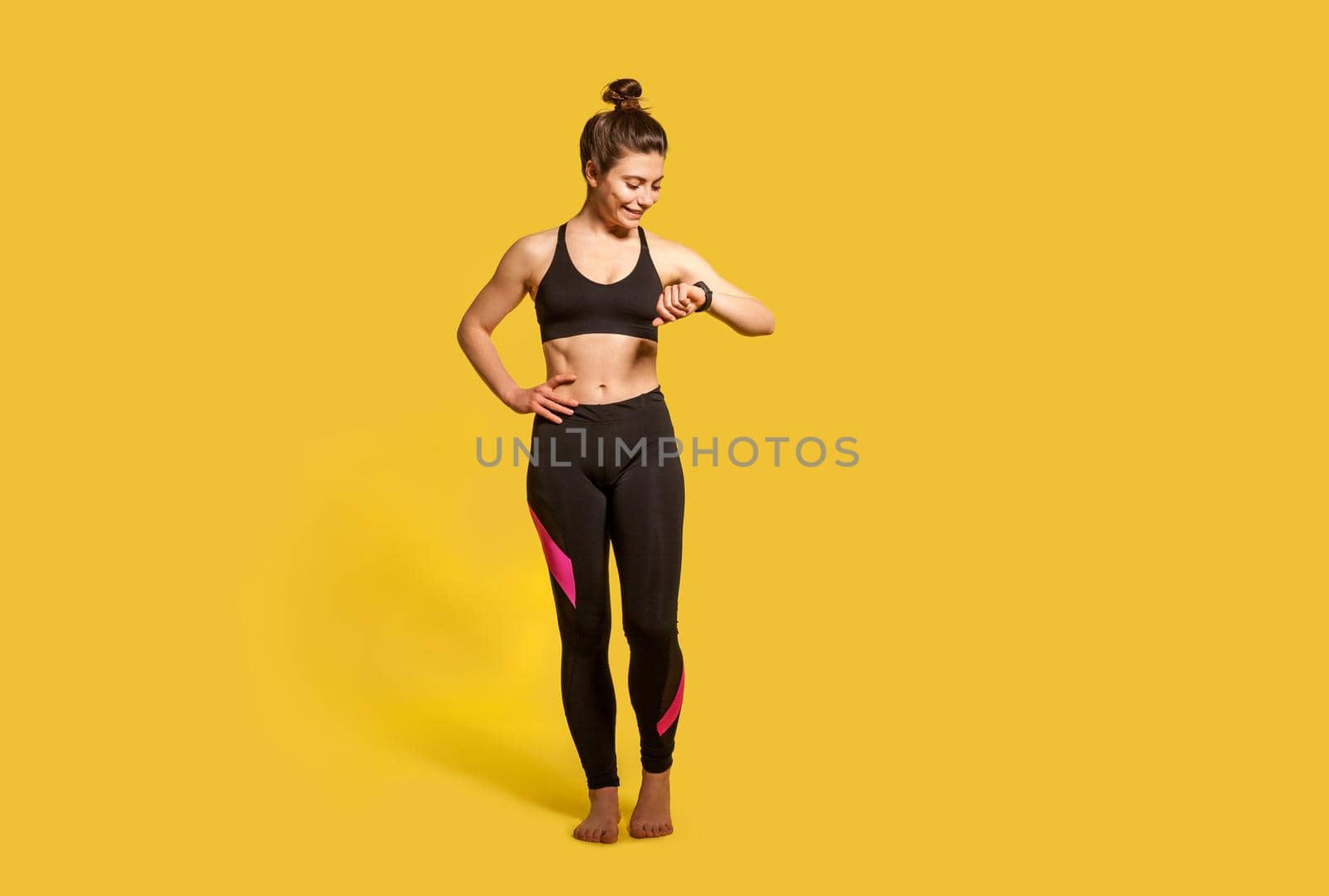 Happy athletic woman with hair bun in tight sportswear looking at wristwatch, checking pulse time on fitness tracker, smiling satisfied with health indicators after trainings. Sport activity, isolated