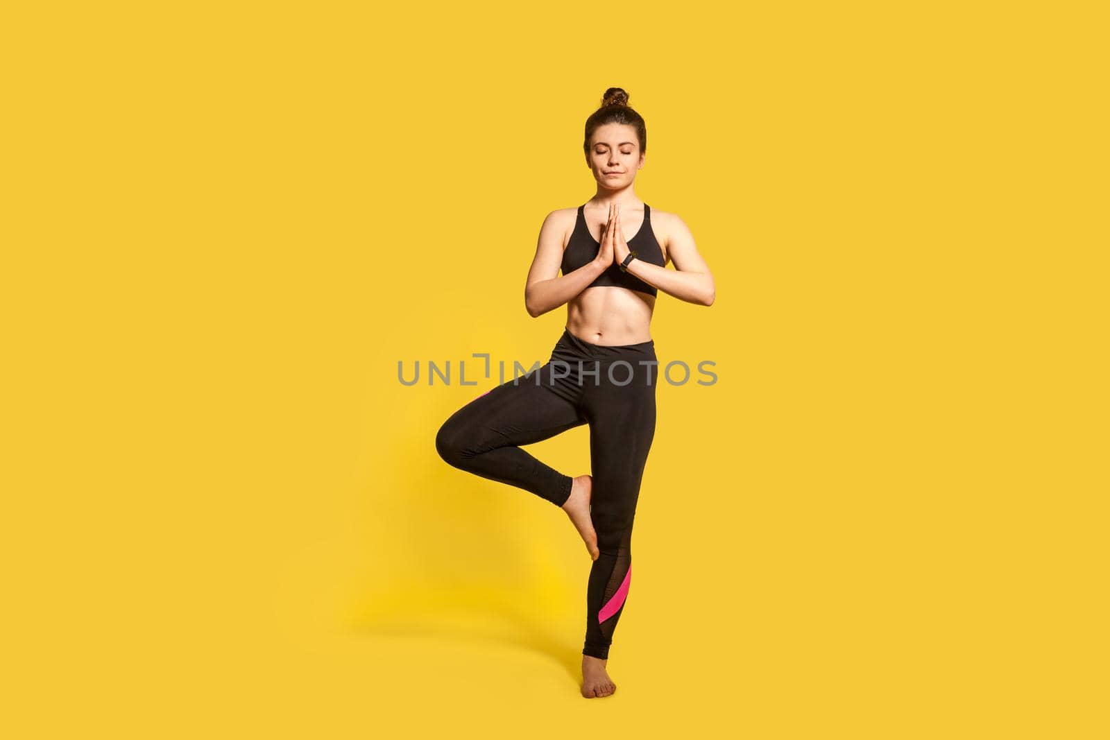 Tree pose. Athletic slim woman with hair bun in tight sportswear practicing yoga, doing Vrksasana exercise on one leg, hands in prayer namaste gesture. full length studio shot, sport workouts isolated