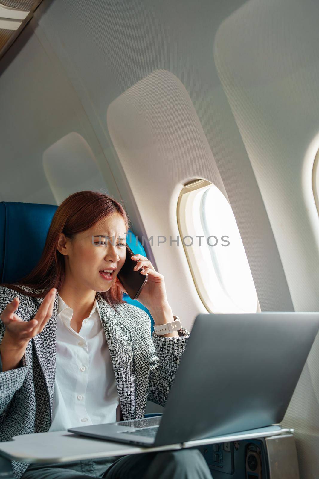Portrait of a successful Asian businesswoman or entrepreneur in a formal suit on an airplane sitting in business class using a phone, computer laptop with a serious expression during the flight by Manastrong
