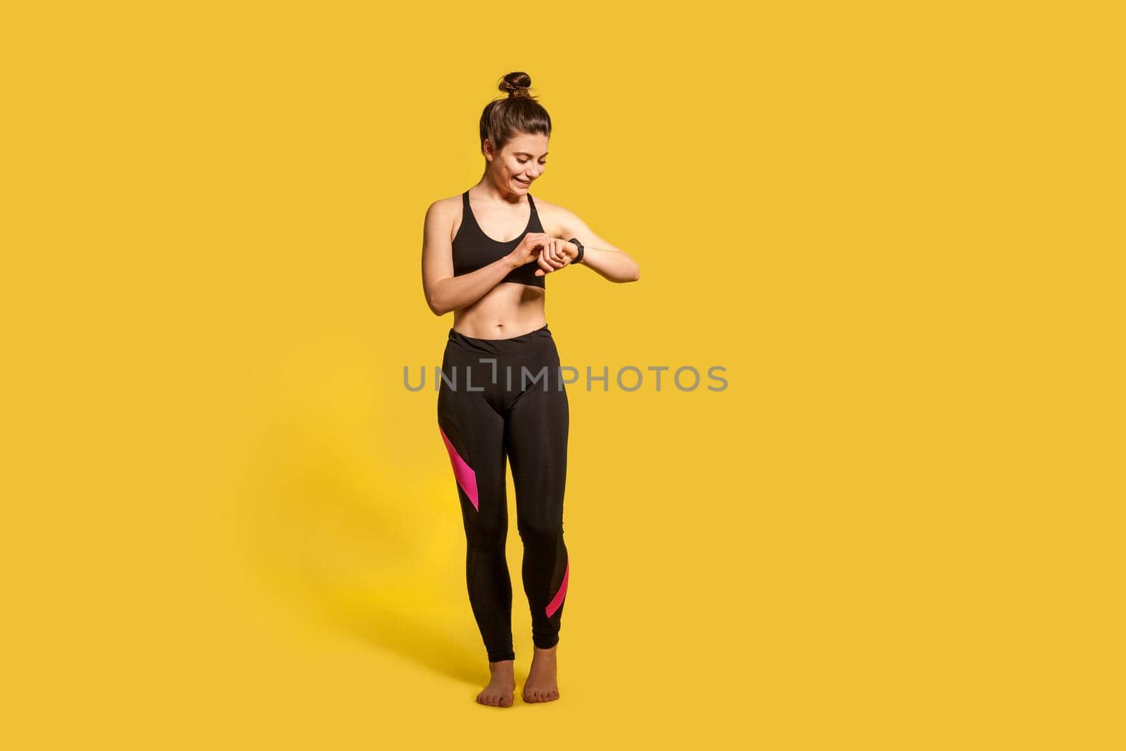 Cheerful athletic woman with hair bun in tight sportswear touching wristwatch display, checking pulse time on fitness tracker, smiling satisfied with health indicators after trainings. Sport activity