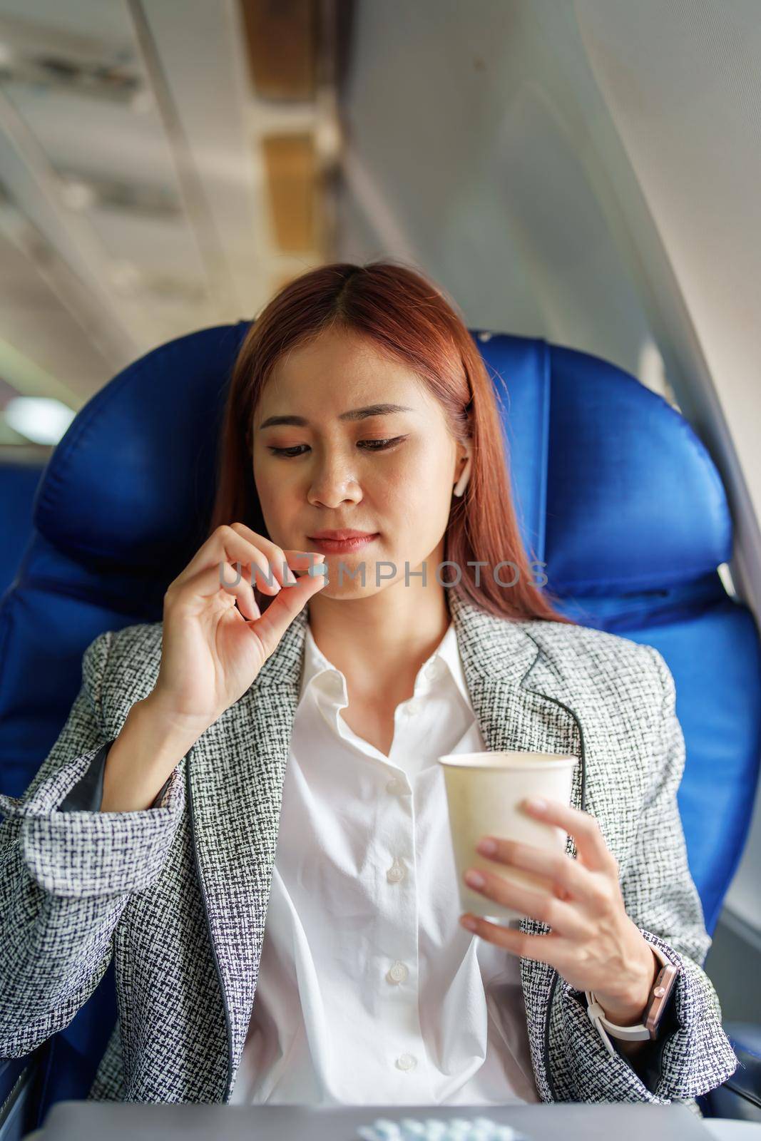 Portrait of a successful Asian businesswoman or entrepreneur in a formal suit on an airplane in business class taking motion sickness pills during the flight by Manastrong