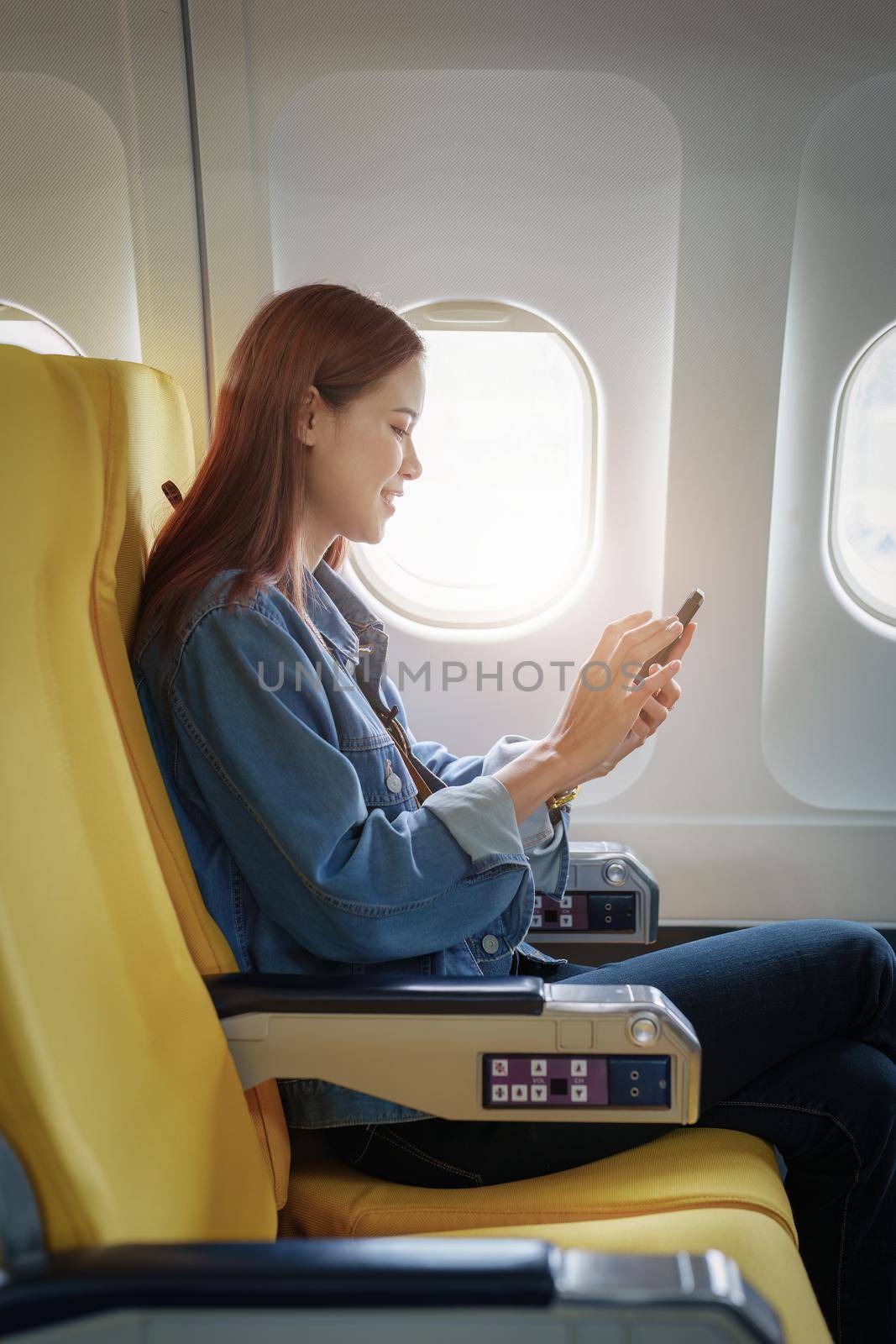 Attractive portrait of Asian woman sitting at window seat in economy class using mobile phone during plane flight, travel concept, vacation.