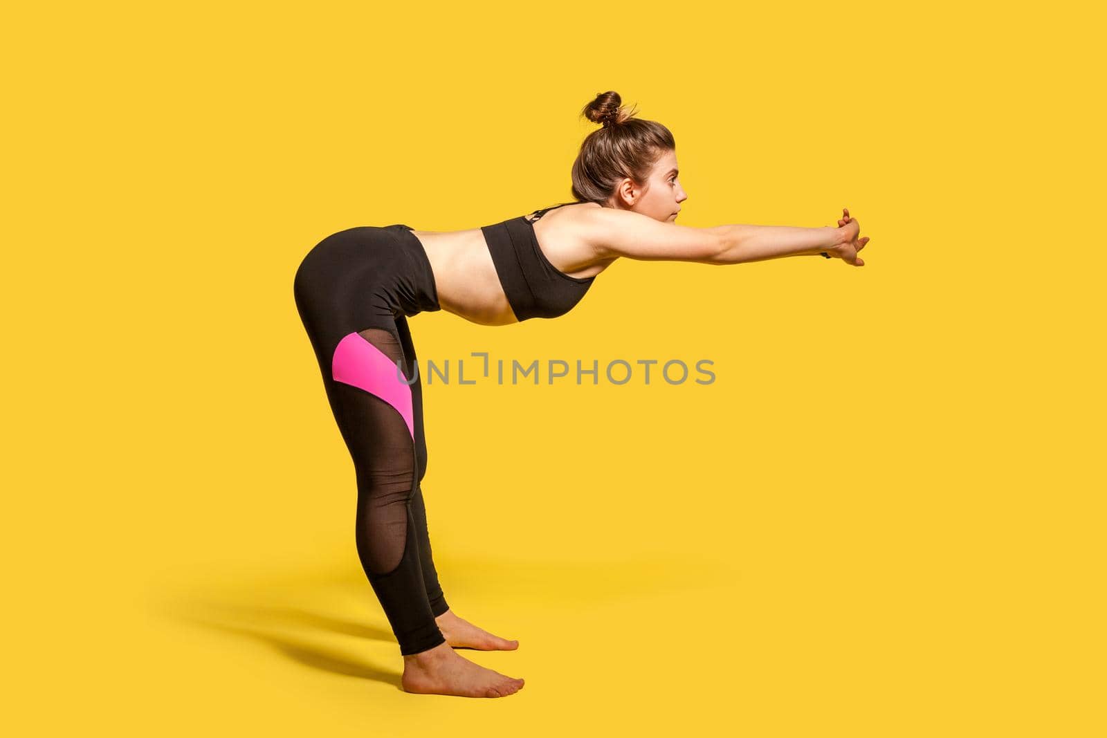 Side view, athletic woman with hair bun in tight sportswear doing sport, bending and stretching hands out, warming up training muscles for flexibility. full length studio shot, isolated on yellow