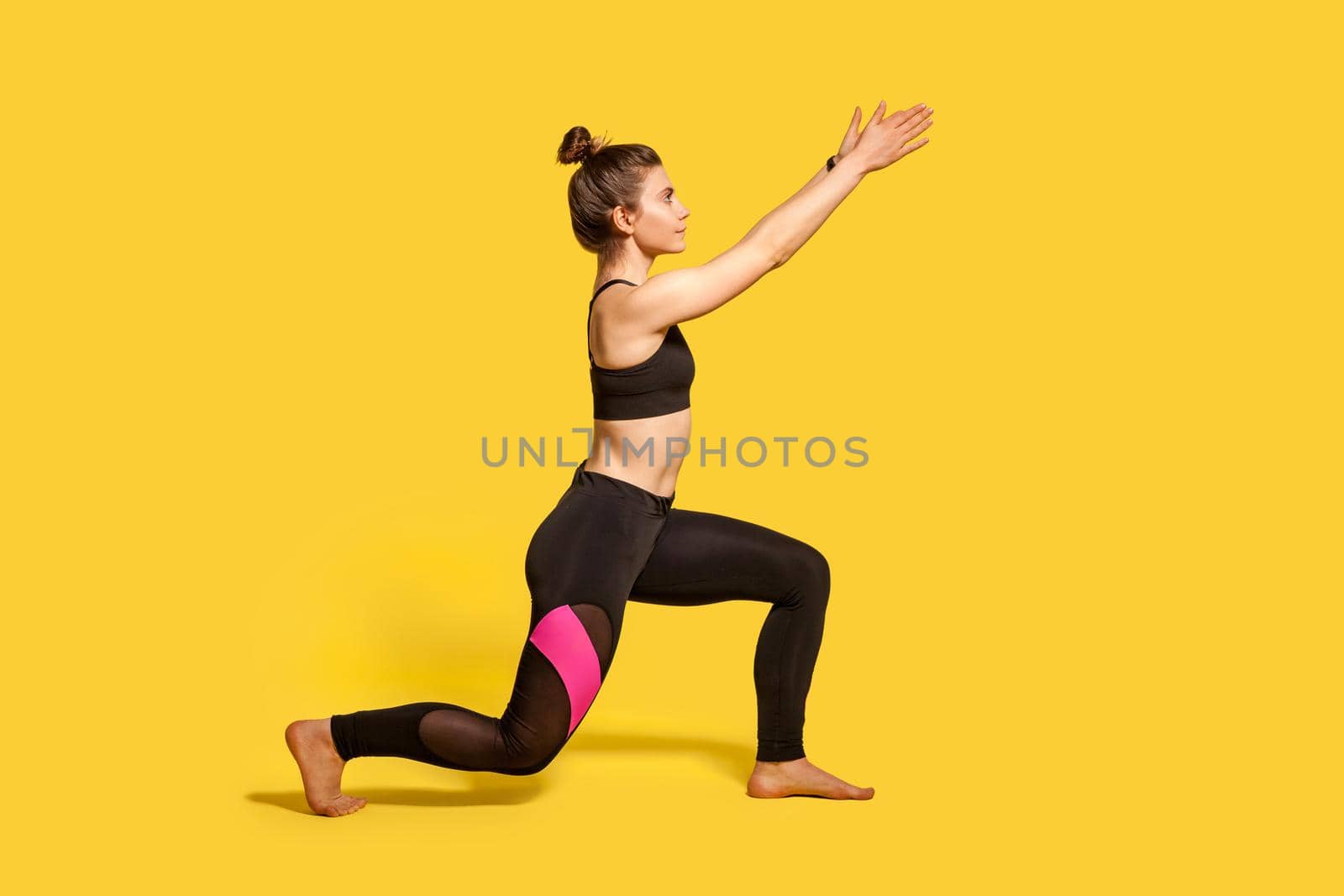 Side view, slim young woman with hair bun in tight sportswear doing sport lunge exercise, standing one knee and raising hands, warming up training muscles. full length studio shot, isolated on yellow