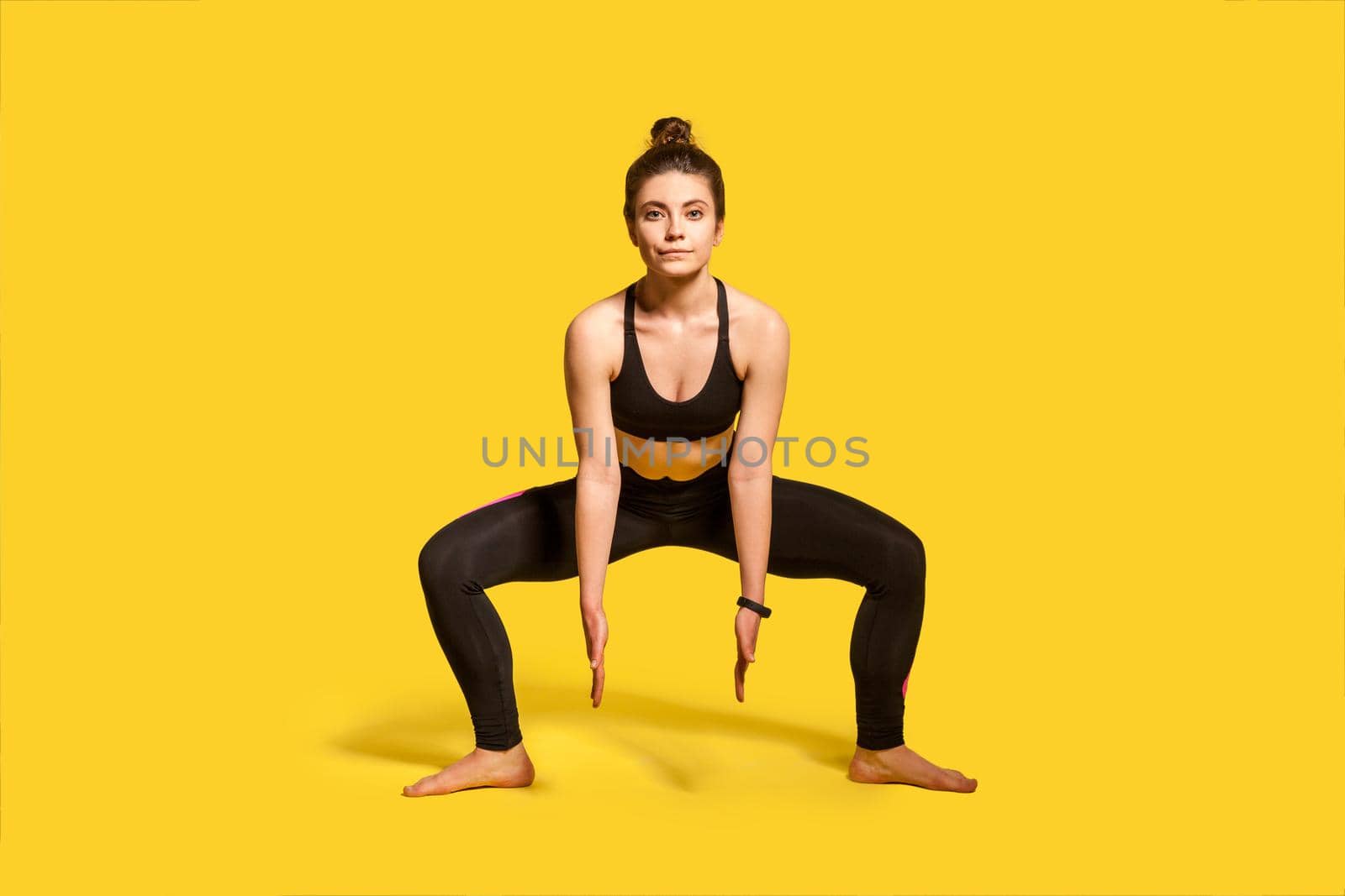 Motivated athletic woman with hair bun in tight sportswear doing frog squat, lower body sport exercise, warming up and training muscles for strength and flexibility. full length studio shot, isolated