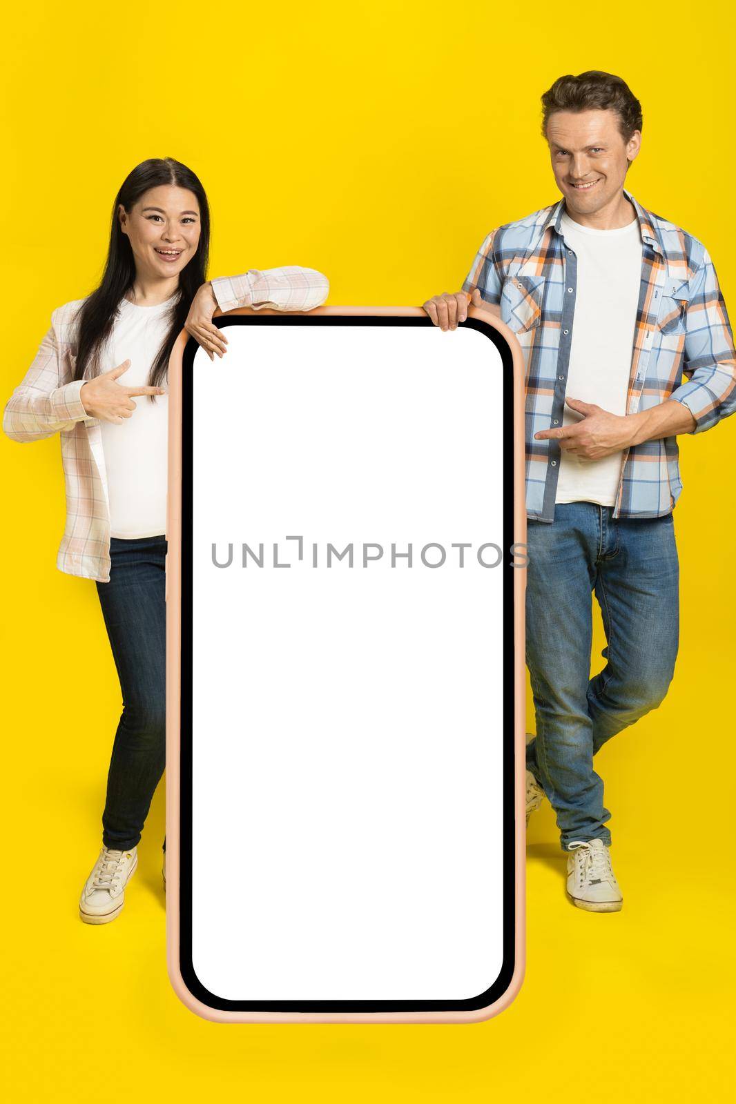 Pointing at white screen caucasian man and asian woman standing leaned on huge smartphone with blank screen, mobile app advertisement isolated on yellow background. Product placement.