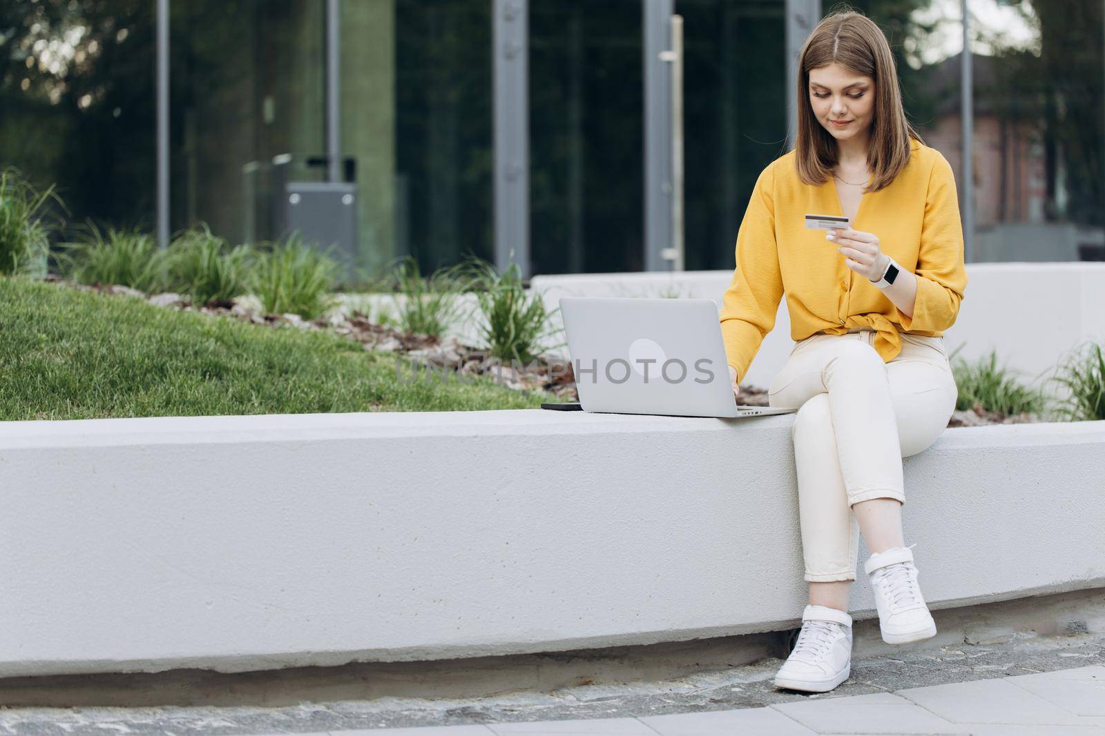 Female shopper using instant easy mobile payments making purchase in online store. E-banking app service. Girl holding credit card and smartphone sitting on bench city buildings outdoor.