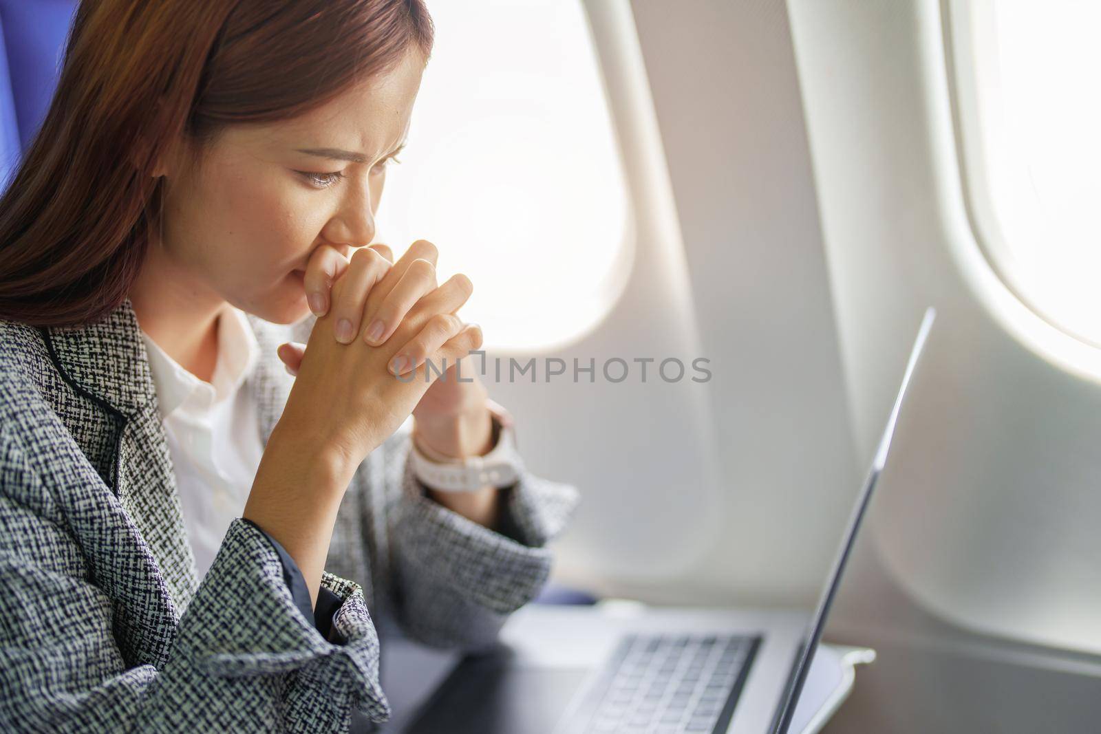 portrait of a successful Asian businesswoman or entrepreneur in a formal suit on an airplane seated in Business Class shows a thoughtful and stressed face with using laptop during the flight.