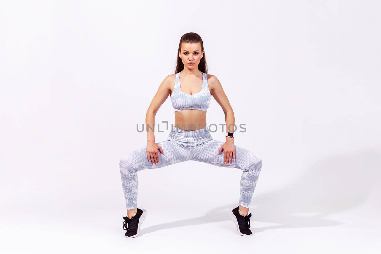 Full length assertive woman in white sportswear squatting and spreading legs to stretch muscles, warming up before twine exercise, training flexibility. Indoor studio shot isolated on gray background