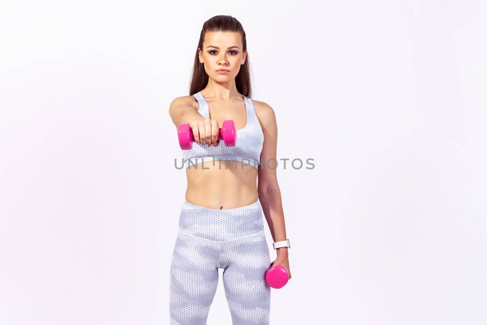 Confident assertive sports woman in white top and tights doing sports exercises holding dumbbells in hands, pumping up muscles, developing strength. Indoor studio shot isolated on gray background