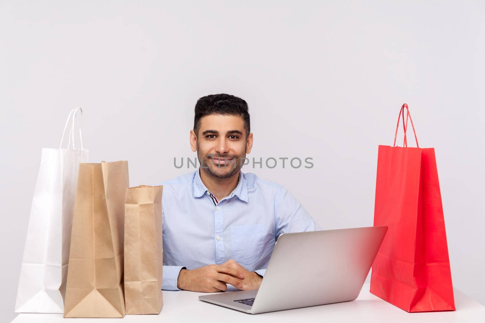 Positive handsome man sitting at desk with laptop, surrounded by packages, looking at camera smiling satisfied with internet shopping, purchase in online store. studio shot isolated, white background