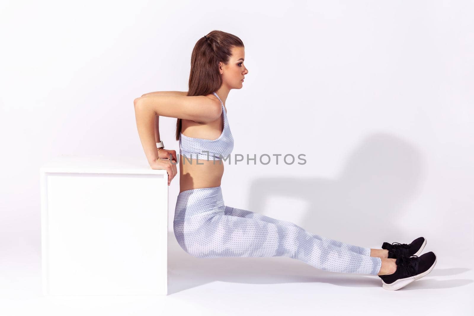 Full length side view assertive sportive woman in white top and tights pumping abs muscles doing sit ups, exercising with cube. Indoor studio shot isolated on gray background