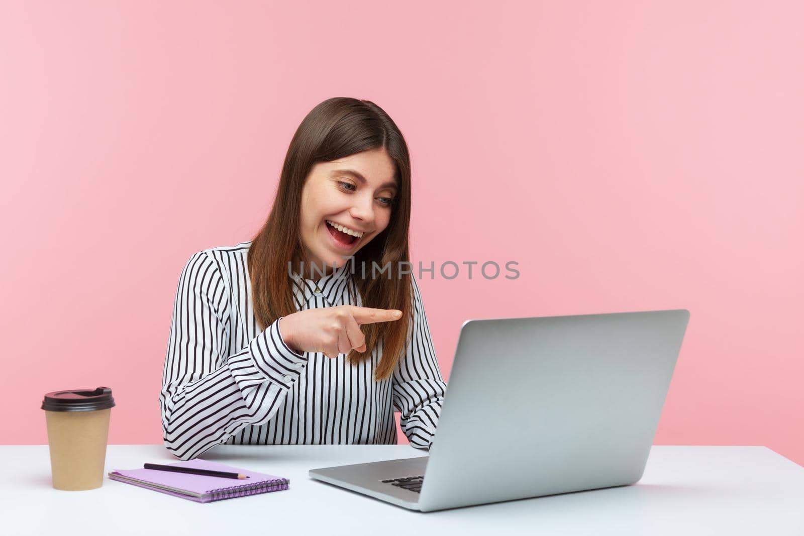 We need you. Positive woman office worker in striped shirt pointing to laptop screen, choosing lucky winner talking on video call, sitting at workplace. Indoor studio shot isolated on pink background