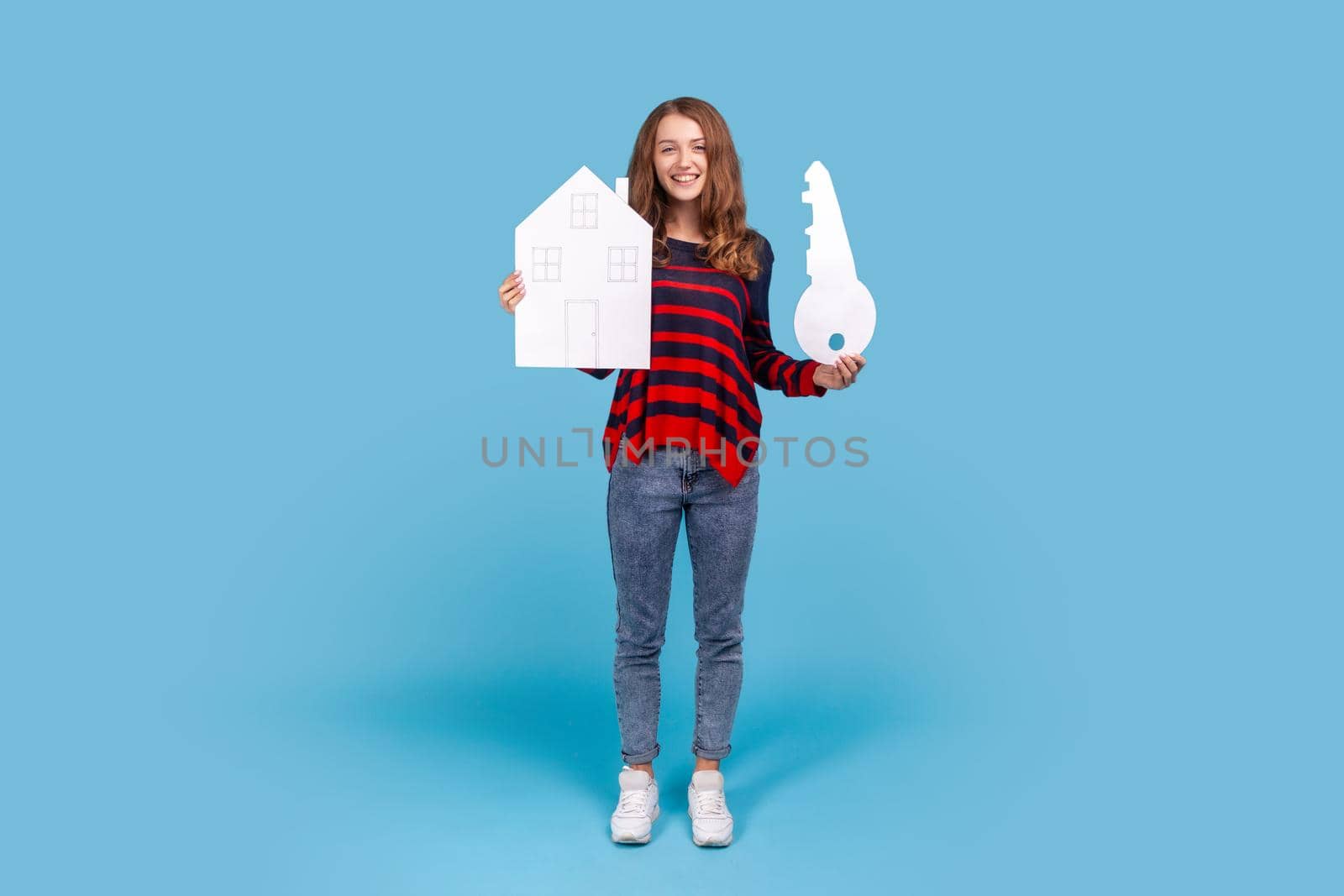 Full length portrait of smiling woman wearing sweater, holds big key and paper house, looking at camera with smile, rejoicing purchase of real estate Indoor studio shot isolated on blue background.