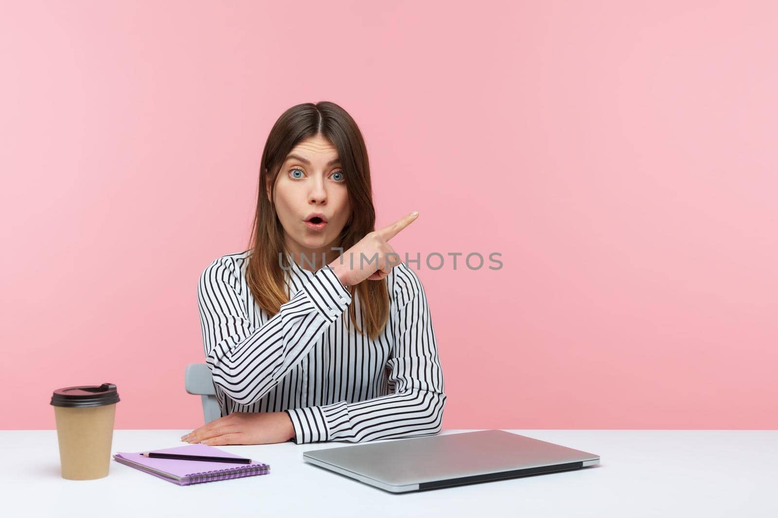 Excited shocked woman pointing finger away paying your attention at empty space for advertising banner, looking at camera with astonishment. Indoor studio shot isolated on pink background