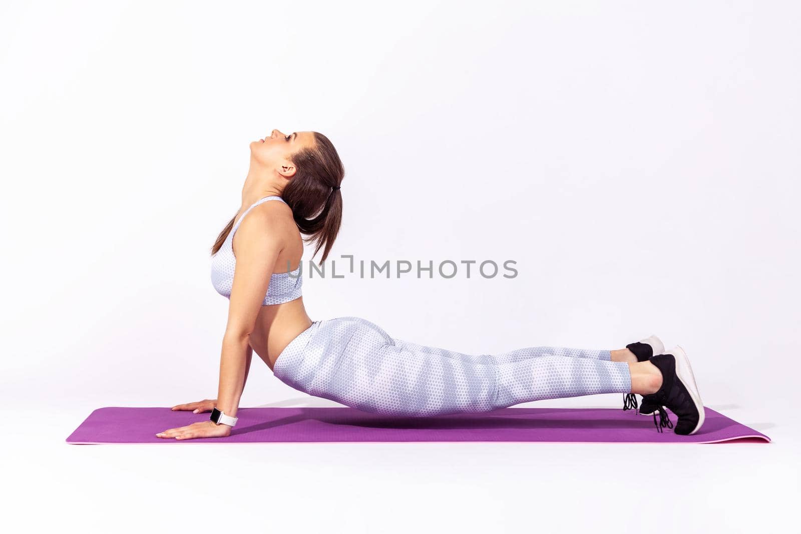Full length athletic woman in white sportswear doing cobra pose, practicing bhujangasana in yoga, stretching back muscles for better flexibility. Indoor studio shot isolated on gray background
