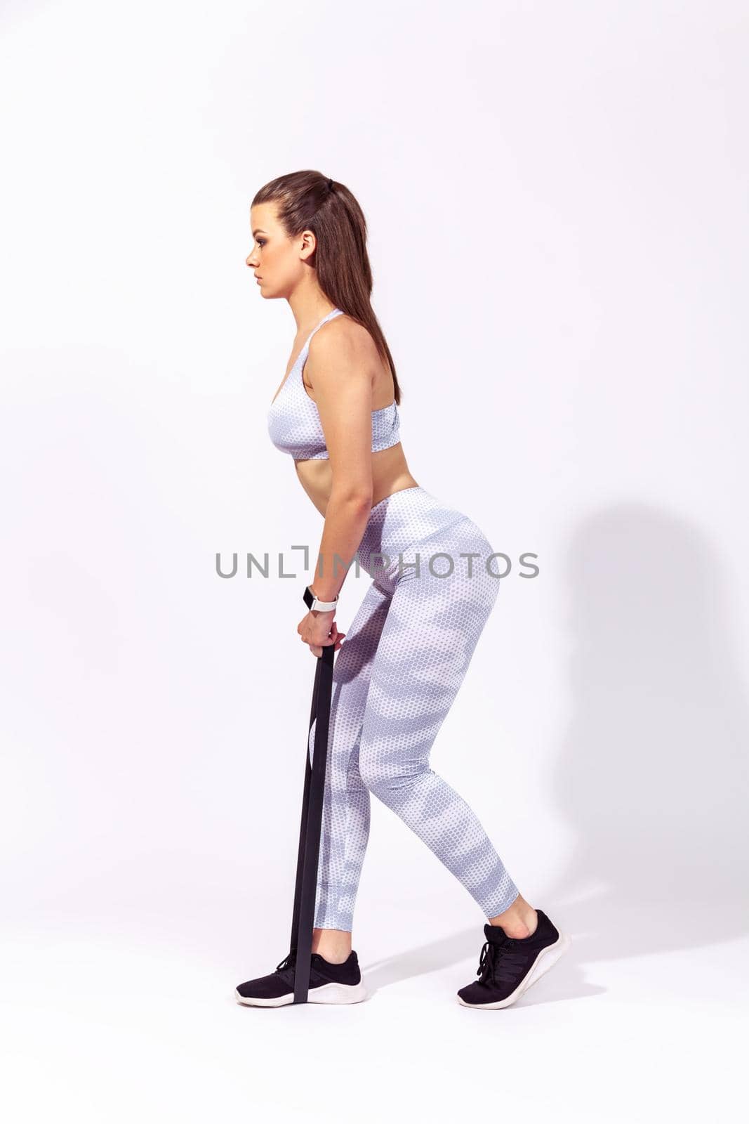 Full length side view motivated athletic woman doing sports, standing on rubber stripe training legs, exercising with rubber expander. Indoor studio shot isolated on gray background