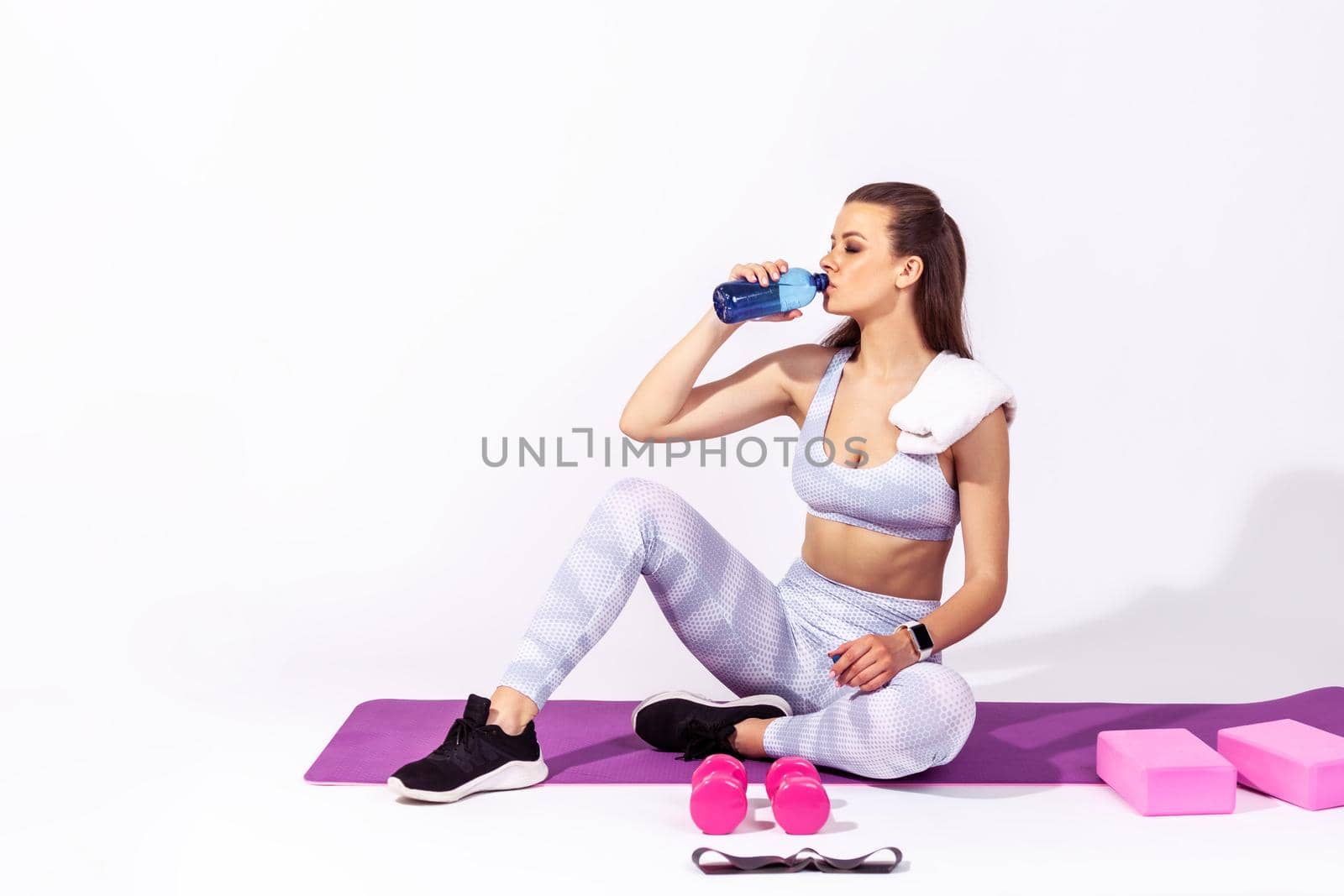 Full length slim athletic woman in white sportswear sitting on fitness mat drinking mineral water, restoring water balance and resting after workout. Indoor studio shot isolated on gray background