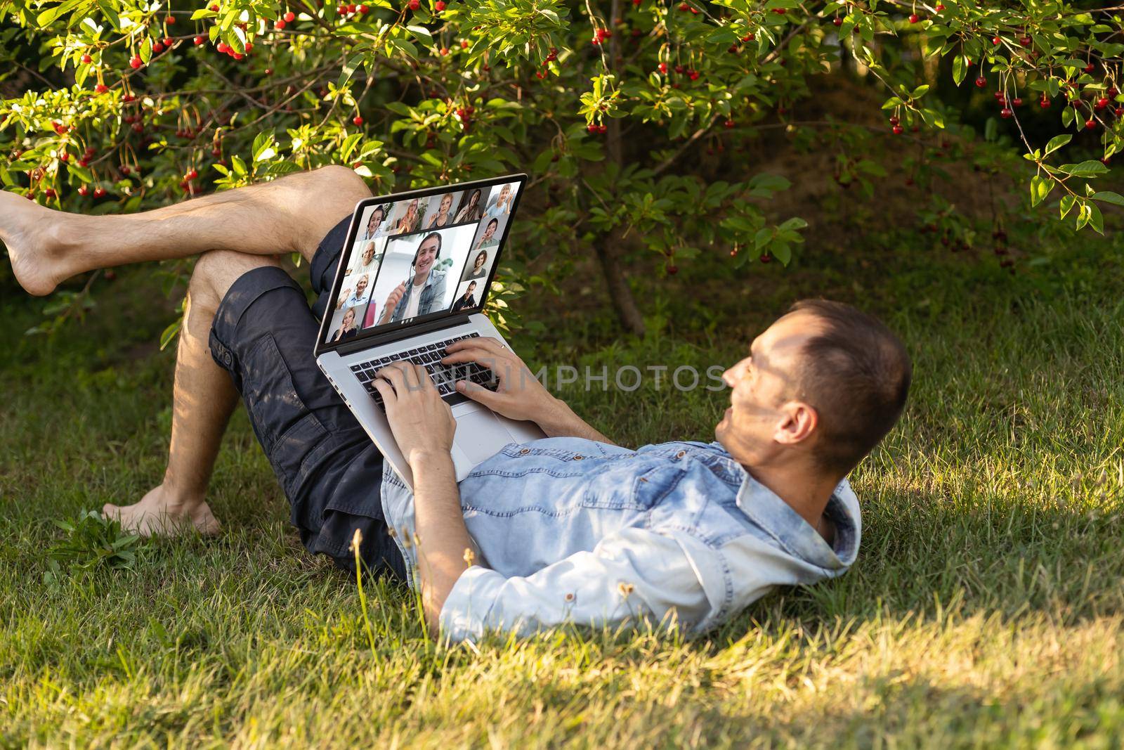 Freelancer man with laptop working in garden sit in chair on grass outdoors. Young blogger male work on computer in public park processes video for social media content. Place of work, distance job