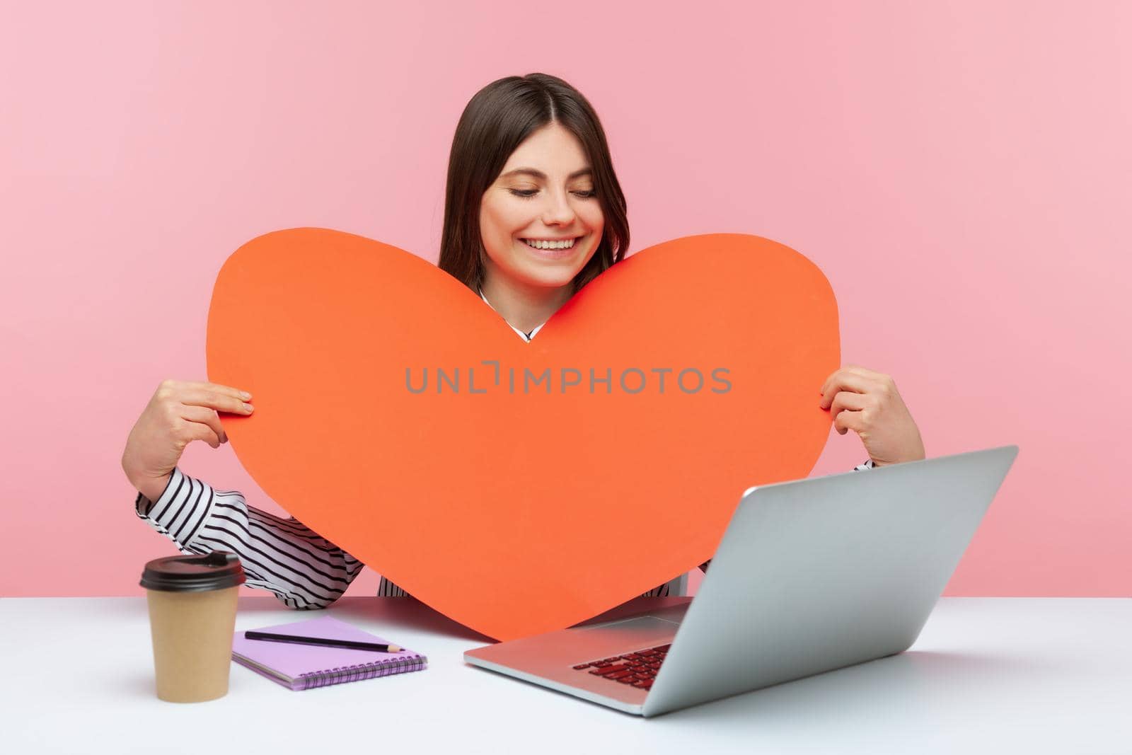 Positive cheerful woman holding big red paper heart talking on video call on laptop sitting at workplace, romantic date online. Indoor studio shot isolated on pink background