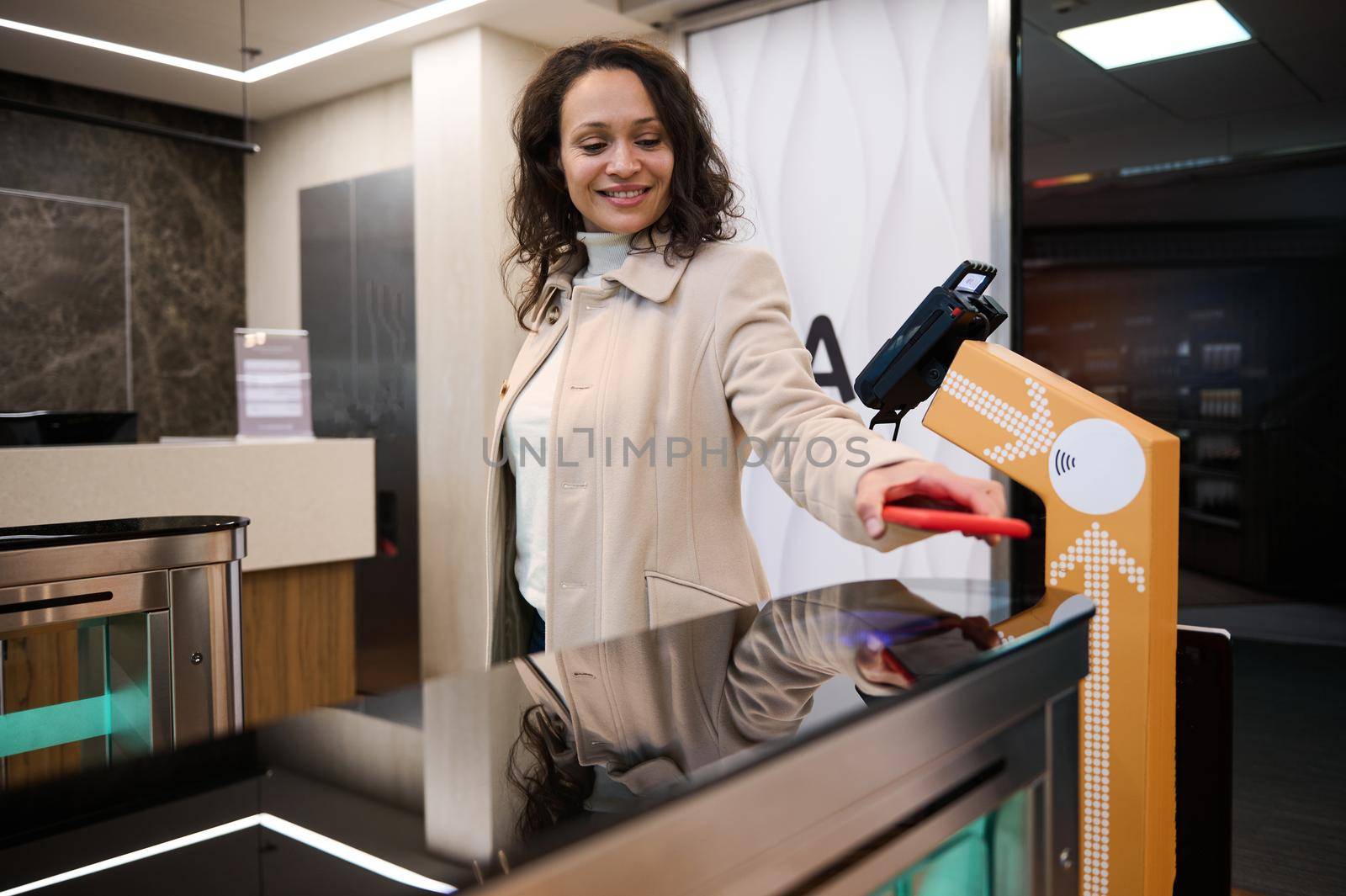 Charming successful Hispanic young woman placing her smartphone over QR code reader at check-in counter while entering to a VIP lounge in the airport departure terminal during her business trip