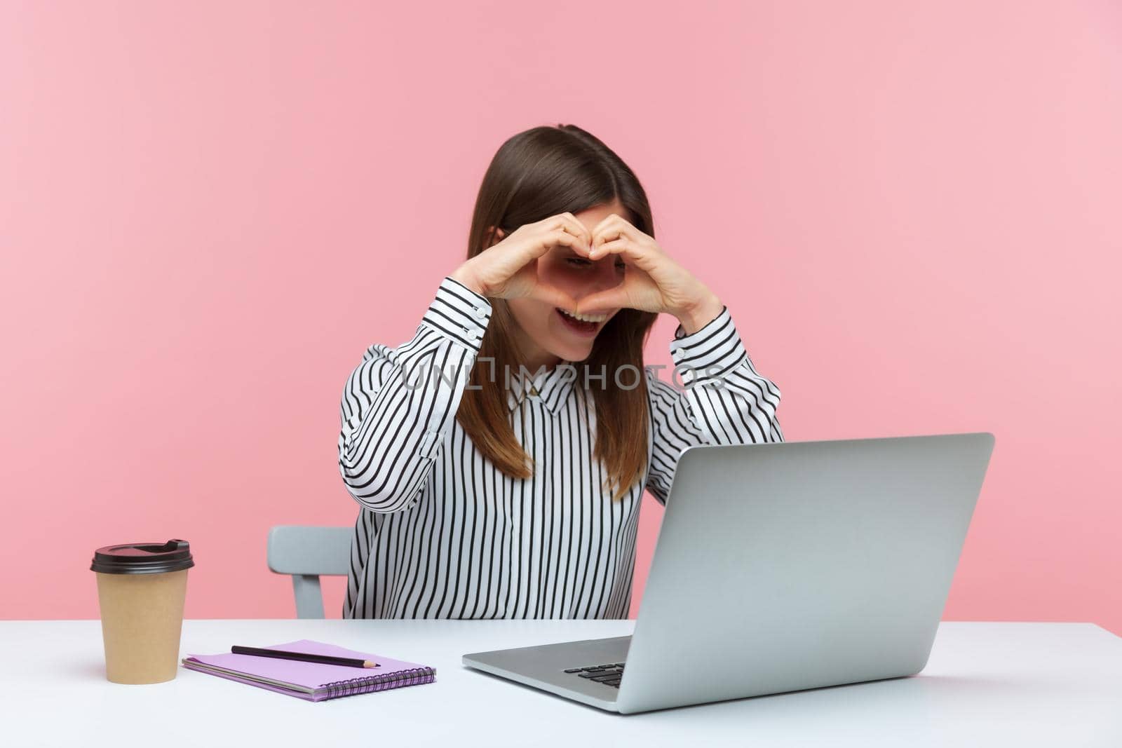 Positive pretty brunette woman office worker making heart with hands, expressing love and looking at laptop screen, talking on video call, online date. Indoor studio shot isolated on pink background